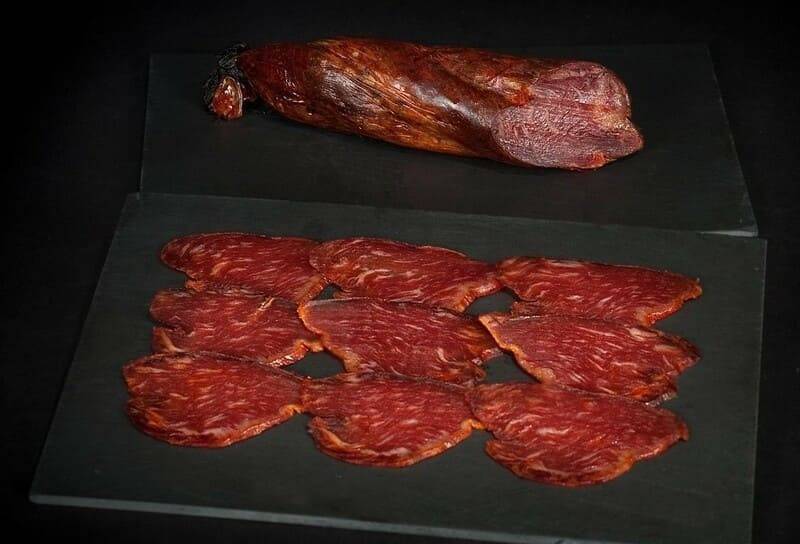 Ganzer Morcón Ibérico de Bellota und aufgereihte Scheiben auf schwarzem Schiefer