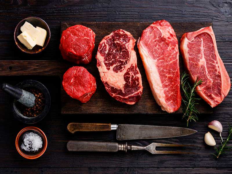 Rohe Rindersteaks und zwei Filetmedaillons auf dunklem Brett mit Tranchiermesser, Gabel, Salz und Butter